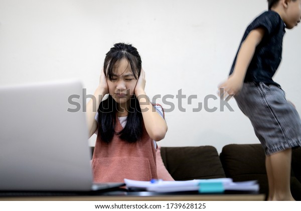 Asian child girl is using her hands to cover her ears\
from the loud voice,annoying noise from little brother while\
studying online,kid boy is jumping,play near her,problems learning\
online at home 