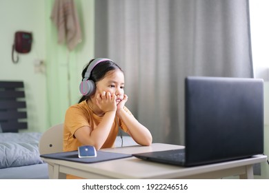 Asian Child Or Kid Girl Bored E-learning On Computer Notebook To Wearing Headphone And Sit To Hand On Chin For Video Call By Unhappy Study Online Or People Learn From Home On Laptop By Back To School