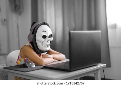 Asian child or kid girl funny e-learning on computer notebook with wearing headphone and white ghost skull mask for video call communication and study online or learn from home school by play laptop 