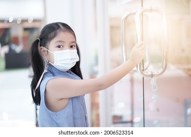 Asian Child Or Kid Girl Holding With Germs Or Bacteria On Glass Door Handle At School Or Mall And Wearing White Face Mask For Close Mouth Nose And Flu To Protect PM 2.5 Dust Or Covid-19 Coronavirus