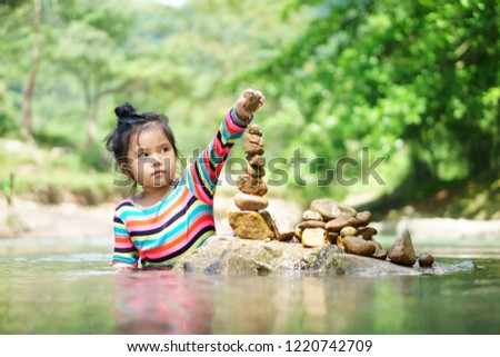 Asian child or kid girl learning sort or stack pebble stone tower or pyramid for water balance training and meditation with nature relax on summer holiday travel at stream or river in forest with tree
