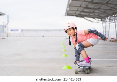 asian child or kid girl playing surf skate or skateboard and crouching with turning practice cones in skating rink or sports park at parking to wearing safety helmet elbow pads wrist and knee support