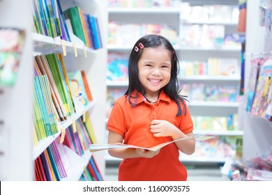 Asian Child Or Kid Girl Red Shirt Happy Smile And Shopping Or Choose Tale Or Story Book And Read On Bookshelf In Bookstore Or Library Room At Kindergarten School Or Nursery For Learn And Study
