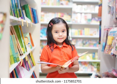 Asian Child Or Kid Girl Red Shirt Happy Smile And Reading Or Choose Tale Or Story Book And Shopping On Bookshelf In Bookstore Or Library Room At Kindergarten School Or Nursery Learn For Summer Study