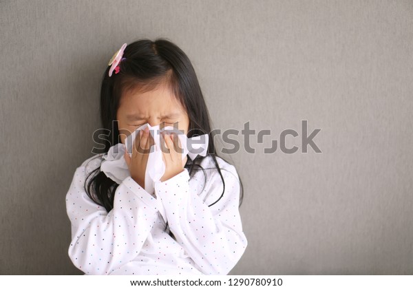 Asian child or kid girl sick with sneezing on nose\
and cold cough on tissue paper because weak or virus and bacteria\
from dust weather and kindergarten and pre school for medical\
background gray space