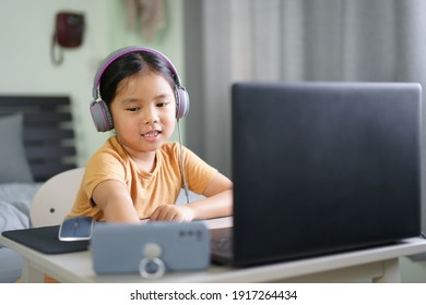 Asian Child Or Kid Girl Smile E-learning On Computer Notebook And Smartphone To Wearing Headphone For Live Streaming Or Video Call By Enjoy Study Online Or People Learn From Home School To Play Laptop