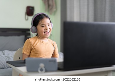 Asian child or kid girl smile learning on computer laptop and smartphone to wearing headphone for live streaming or video call by enjoy study online or people learn from home school to play notebook