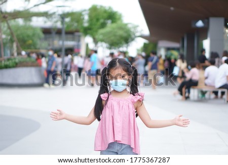 Asian child or kid girl smile wear paper face mask and open arm to present on lot of people for social distancing from person enjoy shopping at department store or market mall on holiday in covid era