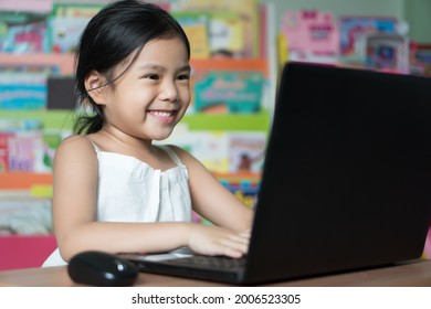 Asian child or kid girl study online or learn from home on computer notebook or people play laptop to communication by primary back to school education and happy smile in library room with bookshelf