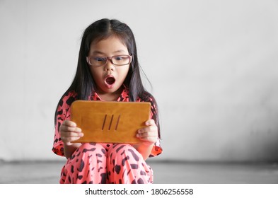 Asian Child Or Kid Girl Wear Blue Block Glasses To Learn From Home Or Study Online On Computer Tablet And Communication With Excited To Open Mouth Wow Or Play Game On Dark Street And Dirty White Wall