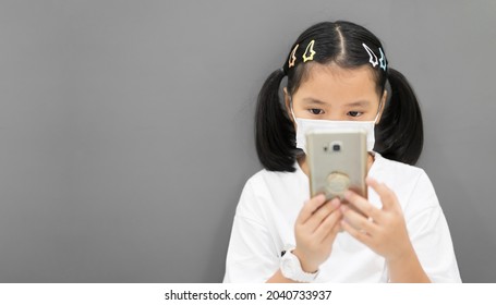 Asian Child Or Kid Girl Wearing White Face Mask Protect Coronavirus And Holding Smartphone To Learn From Home Or Study Online And Communication Or People Play Mobile Phone On Gray Copyspace Background