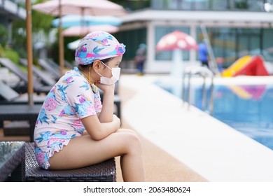 Asian Child Or Kid Girl Wearing Swimsuit And White Face Mask To Protective Coronavirus And Bored Sitting On Bed Or Pool Chair On Swimming Pool And Unhappy In Waterpark For Exercise On Summer Holiday