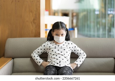 Asian Child Or Kid Girl Wearing White Face Mask To Close Mouth Nose With Flu Sick To Unhappy Angry Sitting Alone On Sofa Chair And Put Hands On Waist For Protect Covid-19 Or Dust At Hospital Or Hotel