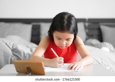 Asian Child Or Kid Girl Write And Read Or Doing Homework On Paper With Thinking Or People Learn From Home And Study Online On Smartphone Or Mobile Phone On Table By Back To School And Wear Red Dress