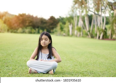 Asian child meditate zen or kid girl smile close eye mind meditating with breath and sit on lawn or meadow grass for peace or yoga relax by hand overlap on tree garden at outdoor park to warm sunshine