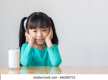 Asian Child Rest Her Chin On Her Hands And Smile Near A Glass Of Milk That Is On The Table