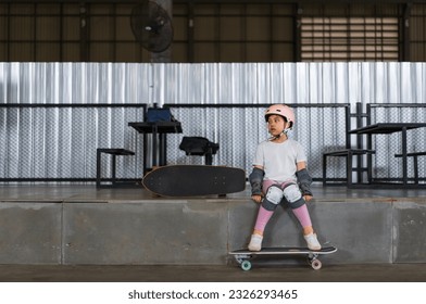 asian child skater or kid girl sitting relax at skatepark with skateboard or surf skate to people extreme sports in indoor surfskate skate park and wears helmet elbow wrist knee guard for body safety