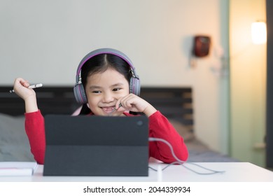 Asian Child Student Back To School Or Kid Girl Smile Learning On Computer Tablet With Holding Touch Pen And Wearing Headset For Video Call Or Person Learn From Home By Study Online Class In Bedroom