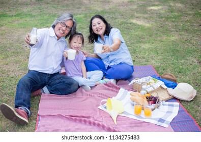 Asian Children And Her Grand Parent Picnic In Garden, They Drinking Milk And Eating  Fruit, Retirement Activity