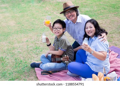 Asian Children And His Grand Parent Picnic In Garden, They Drinking Milk And Eating  Fruit, Retirement Happiness