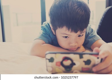 Asian Chinese Boy Playing Smartphone On Bed. Watching Smartphone. Kid Use Phone And Play Game. Child Use Mobile. Addicted Game And Cartoon
