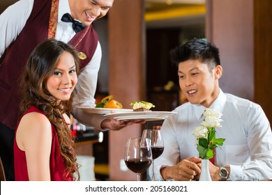 Asian Chinese Couple - Man And Woman - Or Lovers Having A Date Or Romantic Dinner In A Fancy Restaurant While The Waiter Is Serving Food