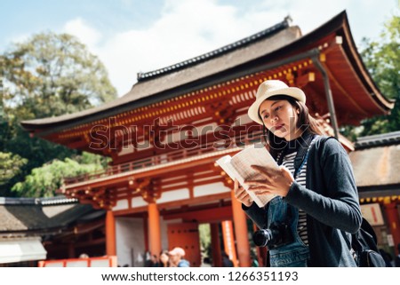 asian chinese female tourist with guide book in grand shrine. young girl reading kasuga taisha history information on sunny day while visiting sightseeing in nara japan. traditional old architecture.