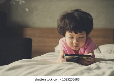 Asian Chinese Girl Playing Smartphone On Bed. Watching Smartphone. Kid Use Phone And Play Game. Child Use Mobile. Addicted Game And Cartoon
