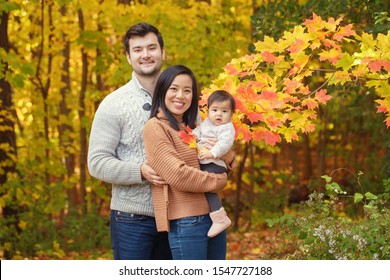 Asian Chinese mother and Caucasian father dad with baby girl in autumn fall park with yellow red tree leaves. Family together outdoor in nature. Seasonal holiday and authentic lifestyle.