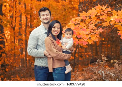 Asian Chinese Mother And Caucasian Father Dad With Baby Girl In Autumn Fall Park. Family Together Outdoor In Nature. Seasonal Holiday And Authentic Lifestyle.