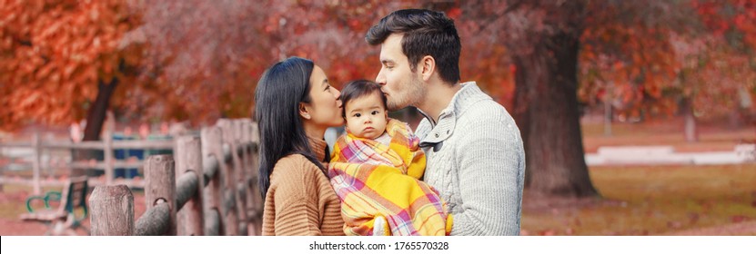 Asian Chinese Mother And Caucasian Father Dad Holding Kissing Baby Girl Wrapped In Blanket. Family In Autumn Fall Park Outdoor. Seasonal Holiday Thanksgiving. Web Banner Header For Website.