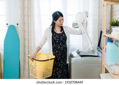 Asian Chinese Pregnant Woman Adding Clothes To Washing Machine And Holding Laundry Basket In Bright Indoors. Young Maternity Wife Putting Husband Shirt Inside. Charming Future Mom Doing Housework.