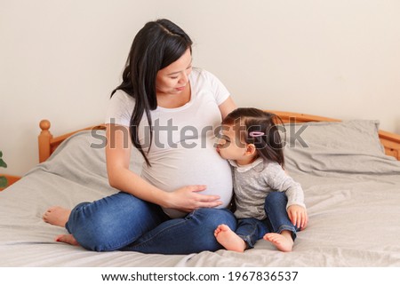 Asian Chinese pregnant woman with toddler girl sitting on bed at home. Girl daughter kid kissing mom belly. Mother and baby daughter expecting waiting for a new family member. Ethnic diversity. 
