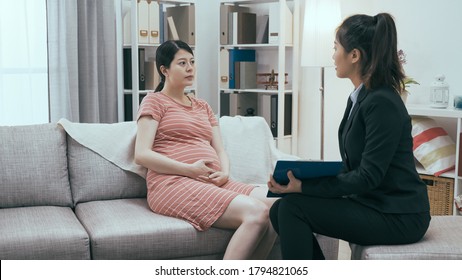 asianische junge chinesische Versicherungspersonal im formalen Anzug bieten eine Lebensversicherung für schwangere Frauen an. Vertriebsmitarbeiterin, die Mutterschaftskundinnen bei einem Besuch zu Hause erläutert.