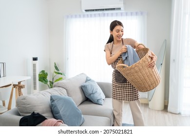 Asian Cleaning Service Woman Worker Cleaning In Living Room At Home. Beautiful Young Girl Housekeeper Cleaner Feel Happy And Take Messy Dirty Clothes Into Basket For Housekeeping Housework Or Chores.