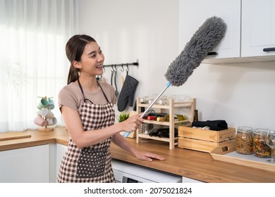 Asian cleaning service woman worker cleaning in kitchen room at home. Beautiful young housekeeper cleaner wear apron, and use feather duster wiping dirty counter for housekeeping housework or chores.