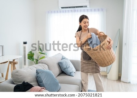 Asian cleaning service woman worker cleaning in living room at home. Beautiful young girl housekeeper cleaner feel happy and take messy dirty clothes into basket for housekeeping housework or chores.