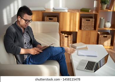 Asian college student sitting on sofa watching some educational program on digital tablet he studying at home - Shutterstock ID 1458115478