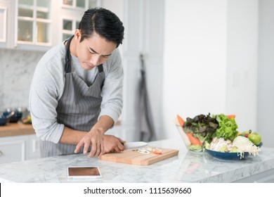 Asian confused man reading his tablet with a pensive thoughtful look while standing in his kitchen while cooking and preparing a meal from a variety of fresh vegetables on the counter
 - Powered by Shutterstock