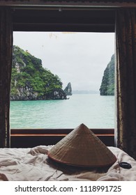 An Asian Conical Hat Rests On A Bed, Looking Out The Window Into Halong Bay, Vietnam From A Cruise Ship Suite.