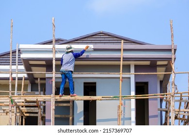 Asian construction worker on wooden scaffolding is painting roof structure of modern house with blue sky background