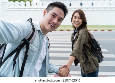 Asian Couple Backpacker Use Mobile Phone Selfie, Take Picture In City. Attractive Man And Woman Tourist Traveler Travel On Street, Using Smartphone Record Vlog On Holiday Vacation Trip In Thailand.