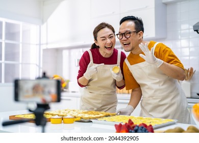 Asian Couple Bakery Shop Owner Using Smartphone With Internet Vlogging Sweet Dessert Baking On Social Media Together In The Kitchen. Small Business Entrepreneur And Online Cooking Class Concept