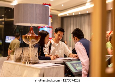 Asian Couple Checking In At Counter With Reception At Hotel.