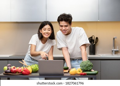 Asian Couple Cooking Together Using Laptop Searching Recipes Online Preparing Dinner Standing In Kitchen At Home. Weekend Leisure, Family Nutrition, Cookery Website Concept. Selective Focus