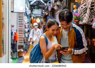 Asian Couple Enjoy And Fun Outdoor Lifestyle Shopping At Street Market On Summer Holiday Vacation. Happy Man And Woman Couple Using Mobile Phone Together While Walking And Shopping At Weekend Market.