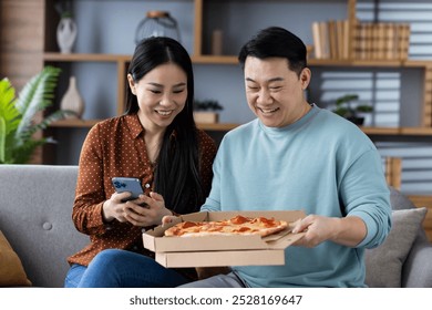 Asian couple enjoying pizza delivery experience, sharing happiness at home. Woman holds phone while man opens pizza box, showcasing joy and togetherness in casual setting. - Powered by Shutterstock