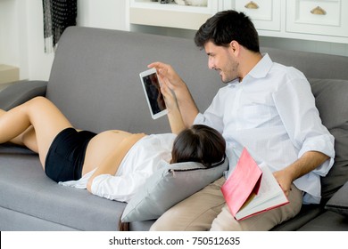 Asian Couple Family Young Mother And Husband Using A Tablet At Home, Woman And Man Is Pregnant Lying On Sofa Looking Tablet Read Book.
