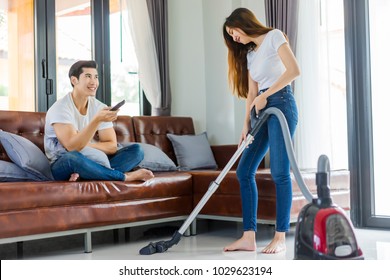 Asian Couple Girl Doing Floor Cleaning With Vaccuum Cleaner While Man On A Brown Sofa At Home Using A Control Remote Watching Television.