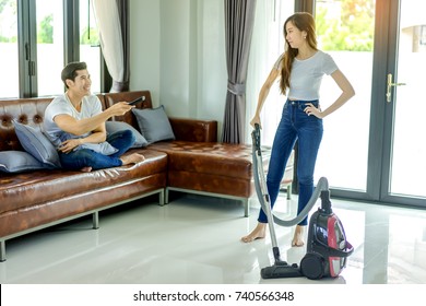 Asian Couple Girl Doing Floor Cleaning With Vaccuum Cleaner While Man On A Brown Sofa At Home Using A Control Remote Watching Television.